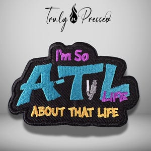 May include: A black embroidered patch featuring the text I'm So A-T-L LIFE ABOUT THAT LIFE. The letters A, T, and L are in bright blue, while the remaining text is in purple and yellow. A small grey graphic is integrated into the letter T.