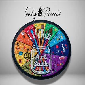 May include: A circular embroidered patch featuring a glass jar filled with various paintbrushes. The background is divided into colorful segments like a color wheel. The text "Art Studio" is embroidered in white on the jar. The brand name "Truly n Pressed" appears above the patch.