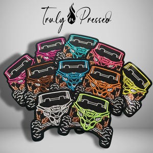 May include: A collection of embroidered patches featuring the front view of off-road vehicles in various neon colors including pink, blue, orange, yellow, white, and green, with brown mud splatter details and the text Truly n Pressed above.