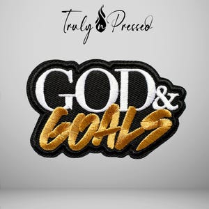 May include: An embroidered patch featuring the text GOD & GOALS in white and gold thread on a black background. The word GOD is in white block letters, while the word GOALS is in gold script. Above the patch, the text Truly Pressed is visible.