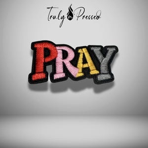 May include: Embroidered patch with the word "PRAY" in red, pink, yellow, and gray letters. The patch has a black border and the text "Truly Pressed" in cursive above the word.