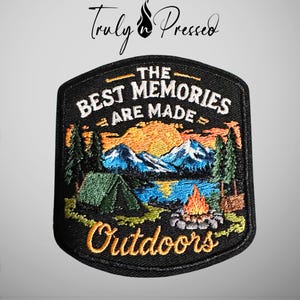 May include: A black embroidered patch featuring a landscape scene with a green tent, a campfire, blue mountains, and a yellow sun. The text reads THE BEST MEMORIES ARE MADE OUTDOORS. The patch has a black border and a shield-like shape.