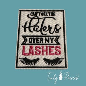 May include: Embroidered patch with the text "CAN'T SEE THE Haters OVER MY LASHES" in black and pink. Features black eyelashes at the bottom. The patch is rectangular with a black border.