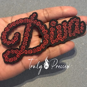 May include: Black embroidered patch in a cursive font spelling "Diva", outlined with red roses. The patch is held in a hand. The text "Truly n Pressed" is written in cursive below the patch.