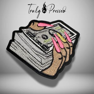 May include: Embroidered patch featuring a stack of money held by a hand with long, pink and gold fingernails. The patch has a black border and the text "Truly Pressed" in cursive script.