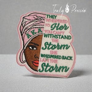 May include: Embroidered patch featuring a woman's face with a pink head wrap and the letters "AKA." The patch includes the text "They whispered to her, you can't withstand the storm. She whispered back, I am the storm."