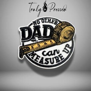 May include: Embroidered patch with the text "NO OTHER DAD CAN MEASURE UP" in black and gold. The patch features a tape measure design and is on a white background. The brand name "Truly Pressed" is at the top.