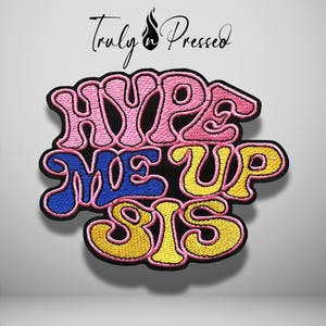 May include: An embroidered patch featuring the text HYPE ME UP SIS in a stylized font. The word HYPE is pink, ME UP is blue, and SIS is yellow. Each word is outlined in black thread, and the entire design is set against a plain background.