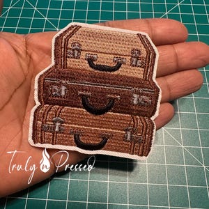 May include: Embroidered patch featuring three stacked vintage suitcases in shades of brown. The patch has a white border and includes the text "Truly n Pressed" in cursive script. The suitcases have black handles and silver-colored clasps.