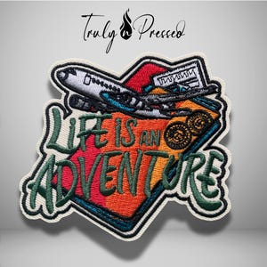 May include: An embroidered patch featuring a white airplane flying over a colorful luggage tag with the text "LIFE IS AN ADVENTURE" in dark green. The tag displays red, orange, and yellow stripes, with two circular brown icons at the top right.