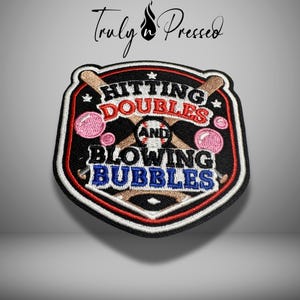May include: A shield-shaped embroidered patch featuring two crossed baseball bats, a baseball, and pink bubblegum bubbles. The text reads HITTING DOUBLES AND BLOWING BUBBLES in red and blue thread against a black background with white and red borders.