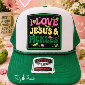 May include: A white and green trucker hat featuring a black rectangular patch with embroidered text that reads I LOVE JESUS & PICKLES in pink, yellow, and green. The design includes small pickle and flower icons, with a green brim and snap closure.