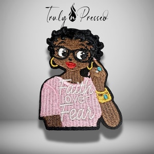 May include: Embroidered patch featuring a cartoon woman with glasses, gold hoop earrings, and a pink sweater. The patch has the words "Faith over Fear" in white script. The woman has brown skin, black hair, and red lipstick. The patch is a unique accessory.