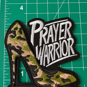 May include: An embroidered patch featuring a high-heeled shoe with a green, black, and tan camouflage pattern, paired with the white stitched text "PRAYER WARRIOR" on a black background, positioned on a green cutting mat with inch markings.
