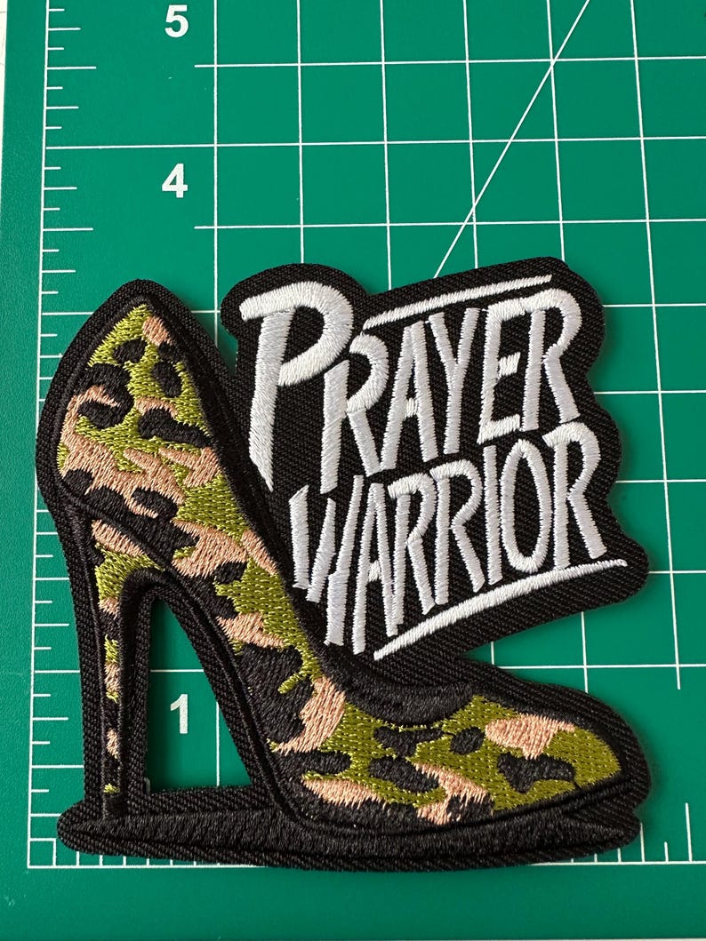 May include: An embroidered patch featuring a high-heeled shoe with a green, black, and tan camouflage pattern, paired with the white stitched text "PRAYER WARRIOR" on a black background, positioned on a green cutting mat with inch markings.
