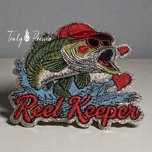 May include: Embroidered patch featuring a cartoon bass wearing sunglasses and a red cap, with red lips and two heart-shaped accents. The text "Reel Keeper" is in red script, with a silver border. The fish is jumping out of blue water.