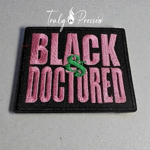 May include: A black embroidered patch with the words "BLACK DOCTORED" in pink thread. A green ampersand symbol is between the words. The patch has a black border and the text "Truly n Pressed" in white cursive.