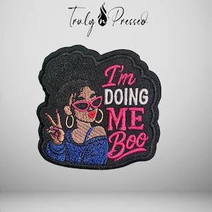 May include: An embroidered patch featuring a person with a high bun hairstyle, wearing pink cat-eye glasses, gold hoop earrings, and a blue top. The person is making a peace sign. The text reads I'm DOING ME Boo in pink and white thread.