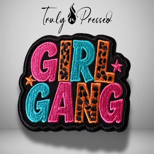 May include: Embroidered patch with the words "GIRL GANG" in pink, teal, and orange, with a leopard print pattern on the letters "R" and "A". The patch has a black border and includes a small star. The text "Truly Pressed" is above the patch.
