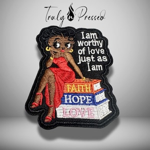 May include: An embroidered patch featuring a cartoon character with black hair in a red dress sitting on a stack of three books labeled Faith, Hope, and Love. The text I am worthy of love just as I am is embroidered in white on the black background.