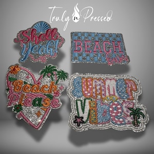 May include: Four embroidered patches with silver glitter backgrounds. Top left: shell shape with pink and blue text Shell yeah!. Top right: rectangular checkerboard with pink text BEACH bum. Bottom left: heart shape with orange text Beach Please. Bottom right: text SUMMER VIBES.