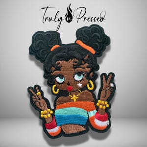 May include: An embroidered patch featuring a cartoon character with dark skin, black hair in two buns, and a colorful striped top. The character is making peace signs with both hands, wearing gold hoop earrings, a star necklace, and beaded bracelets.