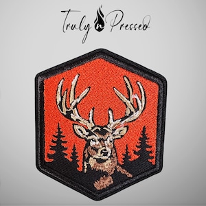 May include: A hexagonal embroidered patch featuring a brown deer head with large antlers centered against a bright orange background. Black silhouettes of pine trees flank the deer, and the entire design is bordered by a thick black stitched edge.