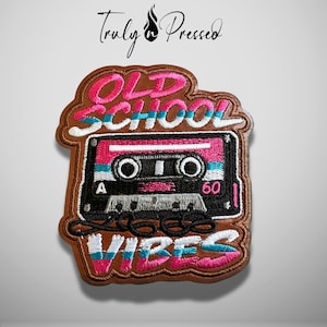 May include: An embroidered patch featuring a black cassette tape with pink and white accents. The text OLD SCHOOL VIBES is stitched in pink and white above and below the tape. The patch has a brown border and a brown background.