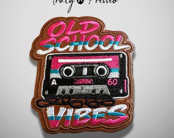 May include: An embroidered patch featuring a black cassette tape with pink and white accents. The text OLD SCHOOL VIBES is stitched in pink and white above and below the tape. The patch has a brown border and a brown background.