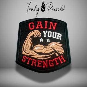 May include: Black embroidered patch with the words "GAIN YOUR STRENGTH" in red and white. The patch features a flexing arm graphic in brown and tan. The patch has a black border and a shield shape.