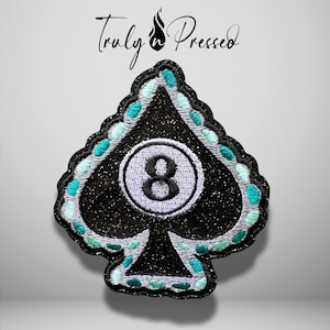 May include: A spade-shaped embroidered patch featuring a black glittery background with a central white circle containing the black number 8. The border is outlined in silver thread with alternating teal and light blue embroidered accents.