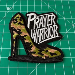 May include: An embroidered patch shaped like a high-heeled shoe with a camouflage pattern in shades of green, tan, and black. The patch features the white embroidered text "PRAYER WARRIOR" on a black background, outlined with a thick black border.