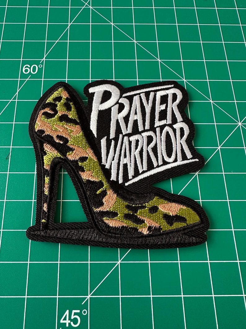 May include: An embroidered patch shaped like a high-heeled shoe with a camouflage pattern in shades of green, tan, and black. The patch features the white embroidered text "PRAYER WARRIOR" on a black background, outlined with a thick black border.
