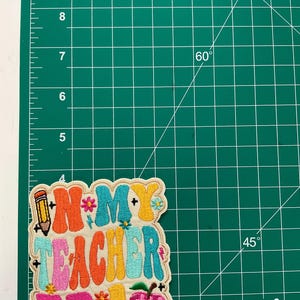 K&ouml;nnte beinhalten: Ein bestickter Aufn&auml;her mit dem Text "IN MY TEACHER ERA" in farbenfroher, wellenf&ouml;rmiger Retro-Schrift. Das Design enth&auml;lt ein Bleistift-Symbol, kleine Blumen und einen karierten Apfel. Der Aufn&auml;her ist etwa 9 cm hoch.
