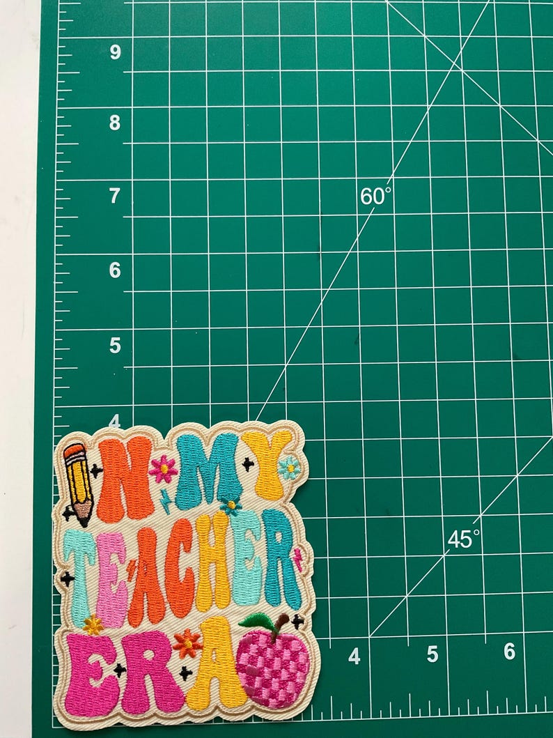K&ouml;nnte beinhalten: Ein bestickter Aufn&auml;her mit dem Text "IN MY TEACHER ERA" in farbenfroher, wellenf&ouml;rmiger Retro-Schrift. Das Design enth&auml;lt ein Bleistift-Symbol, kleine Blumen und einen karierten Apfel. Der Aufn&auml;her ist etwa 9 cm hoch.