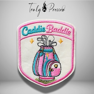May include: A shield-shaped embroidered patch with a white background and a pink border. It features a golf bag with clubs in pink and teal, two yellow stars, and the text "Caddie Baddie" in teal and pink script lettering at the top.
