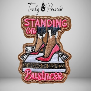 May include: An embroidered patch featuring the text "STANDING ON BUSINESS" in pink thread. The design depicts a pair of legs in dark blue denim and bright pink high-heeled shoes standing on a stack of white paper money with black details.