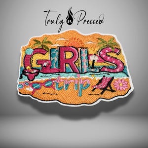 May include: An embroidered patch featuring the text GIRLS trip in pink and blue letters. The design includes a beach scene with palm trees, a sun, a flamingo, pink sunglasses, a beach chair, and a flower on an orange background with a white border.