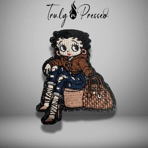 May include: Embroidered patch featuring a cartoon character in a brown jacket and distressed jeans, seated with a handbag. The patch has a black border with the text "Truly Pressed" above the character.