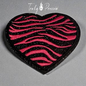 May include: A heart-shaped patch with a black glitter border and a zebra stripe design in bright pink. The text "Truly Pressed" is in white script at the top. This patch is suitable for clothing or accessories.