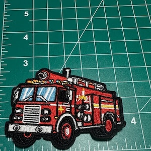May include: Embroidered patch of a red fire truck with black trim. The patch features a detailed design of the fire truck's front, cab, and equipment. The windows are blue, and the wheels are red. The patch is on a green surface.