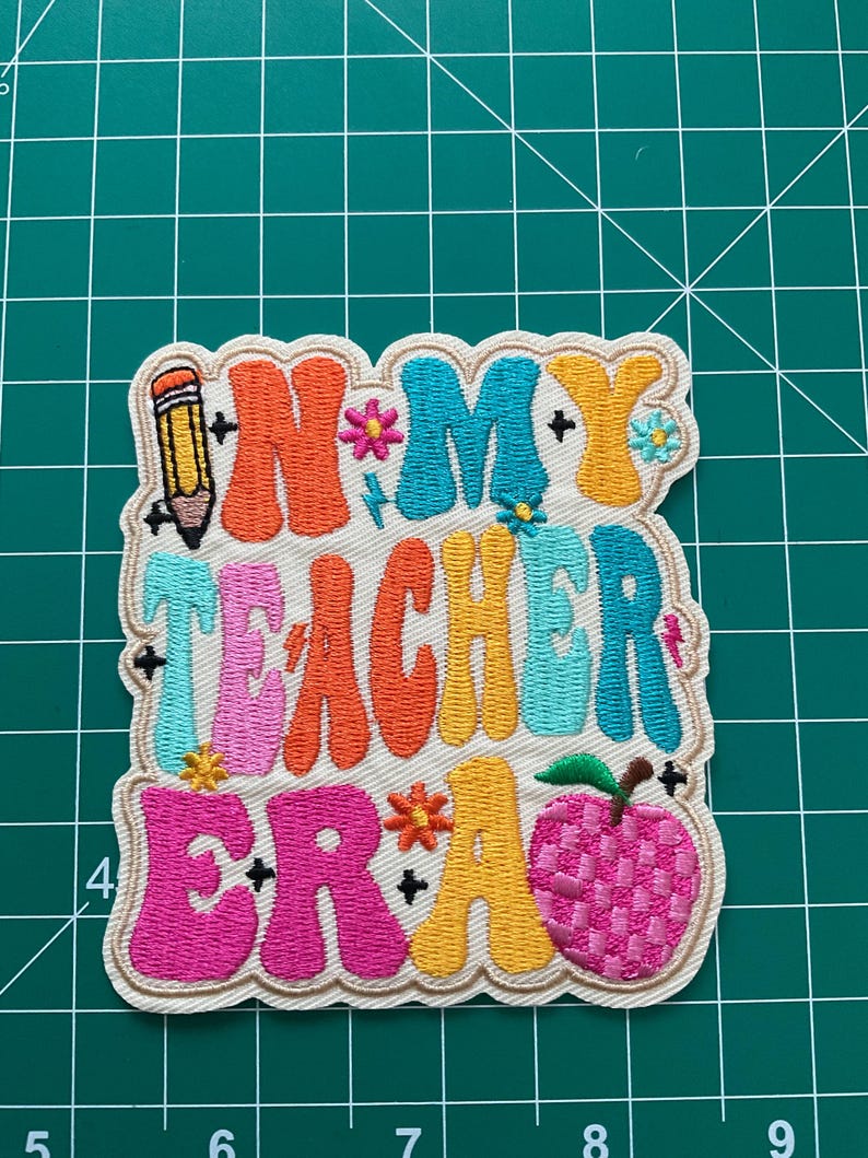 K&ouml;nnte beinhalten: Ein rechteckiger bestickter Aufn&auml;her mit gewelltem Rand, der den Text "IN MY TEACHER ERA" in bunten, wellenf&ouml;rmigen Buchstaben im Bubble-Stil zeigt. Das Design enth&auml;lt einen Bleistift, kleine Blumen und ein rosa Apfelsymbol auf wei&szlig;em Grund.