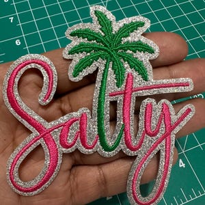 May include: A decorative patch featuring the word Salty in bright pink cursive embroidery with a silver glitter border. A green embroidered palm tree is positioned above the letter l, integrated into the design. The patch measures approximately 4 inches in height.