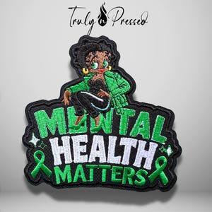May include: An embroidered patch featuring a cartoon character with dark hair and gold earrings wearing a green jacket. The text MENTAL HEALTH MATTERS is stitched in green and white. Two green awareness ribbons are on the sides of the text.