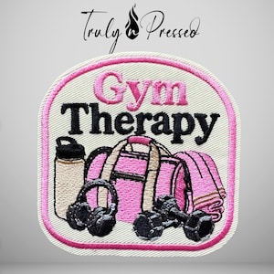 May include: An embroidered patch with a white background and pink border featuring the text Gym Therapy in pink and black. The design includes a beige water bottle, a pink gym bag, black headphones, and a pair of black dumbbells.