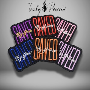 May include: Embroidered patches in various colors, including purple, red, orange, blue, and pink. Each patch features the word "SAVED" and "By Grace" in a contrasting color. The patches are black with a black border. The image also includes the text "Truly n Pressed".