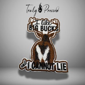 May include: An embroidered patch featuring a brown deer viewed from behind with white tail markings and large antlers. The text reads I like BIG BUCKS and I CANNOT LIE in black lettering. The patch has a white border and a brown stitched edge.