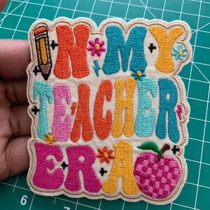 K&ouml;nnte beinhalten: Ein bestickter Aufn&auml;her mit cremefarbenem Hintergrund und dem Text "IN MY TEACHER ERA" in bunten, wellenf&ouml;rmigen Retro-Buchstaben. Das Design enth&auml;lt einen gelben Bleistift, kleine Blumen und ein rosa kariertes Apfelsymbol.