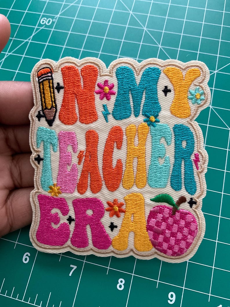 K&ouml;nnte beinhalten: Ein bestickter Aufn&auml;her mit cremefarbenem Hintergrund und dem Text "IN MY TEACHER ERA" in bunten, wellenf&ouml;rmigen Retro-Buchstaben. Das Design enth&auml;lt einen gelben Bleistift, kleine Blumen und ein rosa kariertes Apfelsymbol.