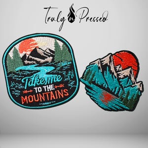May include: Two embroidered patches featuring mountain landscapes. The left patch is oval with a red sun, green trees, blue water, and the text "Take me TO THE MOUNTAINS". The right patch is shaped like a mountain peak with a red sun and blue water.